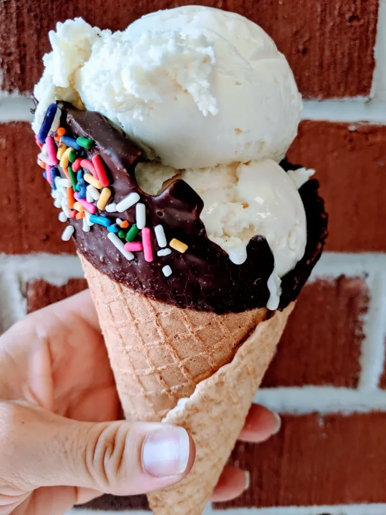 Vanilla scoop in a chocolate and sprinkle dipped waffle cone held against a brick wall